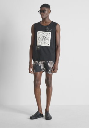 Muškarac obučen u crnu majicu bez rukava s bijelim uzorkom, cvjetne kratke hlače, crne cipele na navlačenje i sunčane naočale, stoji na bijeloj pozadini.