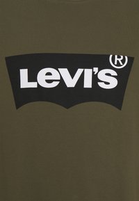 Logo Levi's v bílém textu uvnitř černého zakřiveného obdélníku na olivově zeleném textilním pozadí.