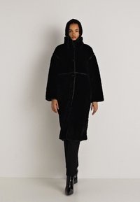 Abrigo de piel sintética negra con cuello alto, corte oversized y grandes botones. Presenta una textura suave y detalles en las costuras laterales.