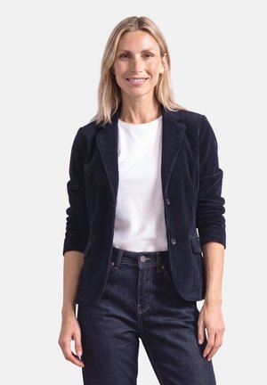 Smilende kvinde med blondt hår iført navy blød fløjlsjakke, hvid skjorte og mørke jeans, stående mod en ensfarvet lys baggrund.