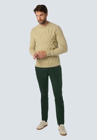 Crème-kleurige gebreide sweater met een golfpatroon, gecombineerd met donkergroene slim-fit broek en lichtbeige sneakers.