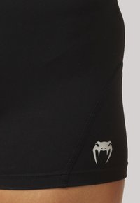 Μαύρες αθλητικές βερμούδες με λεία υφή έχουν το ασημένιο λογότυπο ενός φιδιού στην κάτω πλευρά και ορατές λεπτομέρειες ραφής.