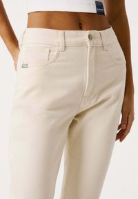Pantaloni in denim beige chiaro con design a gamba dritta, dotati di chiusura frontale con bottone, cinque tasche e una piccola etichetta con il marchio sulla tasca.
