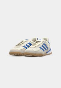 Atletische schoenen met een beige suède bovenkant, blauwe accenten, drie strepen en een gumrubberen zool. Het ontwerp heeft een gewatteerde kraag en veters.