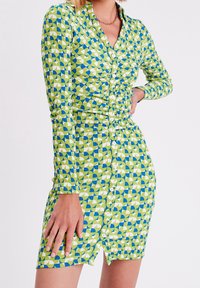 Robe chemise à motifs verts et bleus avec manches longues, fermeture à boutons et détails froncés à la taille. Tissu texturé avec des formes géométriques.