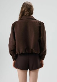 Bruine satijnen bomberjack met kraag, gerimpelde tailleband en lange mouwen, gecombineerd met bijpassende bruine shorts. Soepele textuur, minimale details.