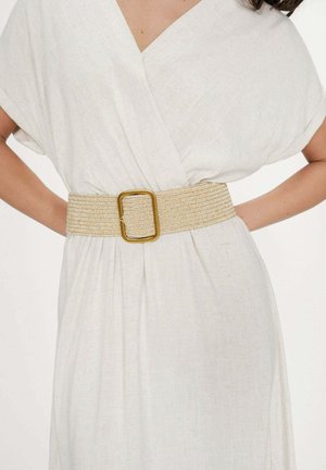 Femme portant une robe blanche à col en V avec des manches courtes, cintrée à la taille par une large ceinture tressée beige avec une grande boucle rectangulaire dorée.