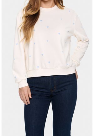 Kobieta ubrana w białą bluzę z małymi niebieskimi sercami oraz ciemnoniebieskie dżinsy, stojąca z jedną ręką na biodrze.