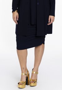 Marineblaues Kleid mit figurbetontem Rock, darüber eine lange, lockere Jacke. Getragen mit goldenen Plateausandalen mit offener Zehe und Knöchelriemen.