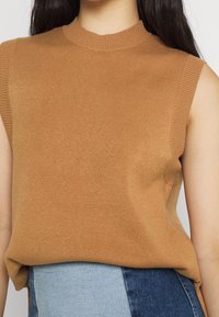 Beżowy top bez rękawów z miękkiego dzianinowego materiału, z prążkowanym, okrągłym dekoltem i luźnym krojem. Dolne krawędzie są lekko asymetryczne.