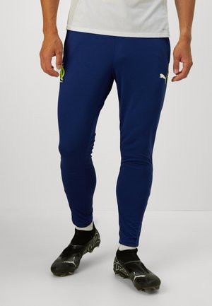 Blauwe sportbroek van vochtafvoerende stof, met een slanke pasvorm, geborduurd geel logo en geribbelde manchetten. Zwarte voetbalschoenen eronder.