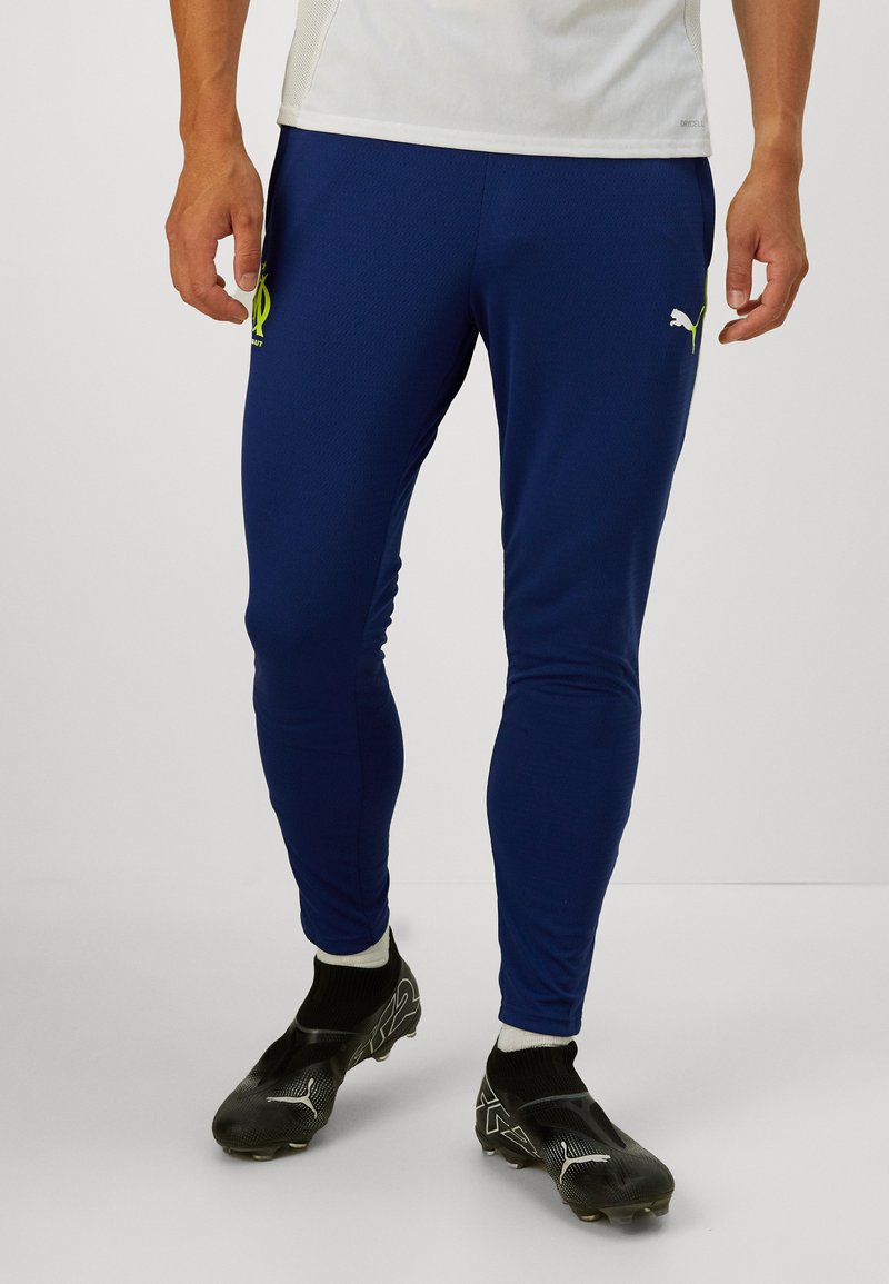 Μπλε αθλητικά παντελόνια από ύφασμα που αποβάλλει την υγρασία, με στενή εφαρμογή, κεντημένο κίτρινο λογότυπο και ραμμένα μανίκια. Μαύρα ποδοσφαιρικά παπούτσια από κάτω.