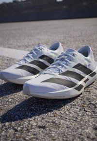 Paar weiße Laufschuhe mit schwarzen Streifen auf Asphalt nahe einer weißen Linie, gezeigt bei Tageslicht im Freien.