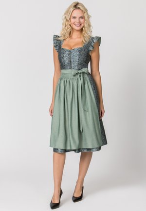 Blonde Frau lächelt, trägt ein blaues Blumendirndl mit Rüschenärmeln und eine hellgrüne Schürze, die in der Taille gebunden ist, schwarze High Heels.