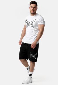 Bijela majica s crnim logotipom "TAPOUT", uparen s crnim kratkim hlačama koje imaju isti logotip. Nosite uz bijele tenisice i prugaste čarape.