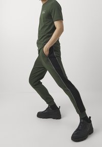 Zelené tričko s krátkým rukávem a logem, doplněné zelenými a černými tepláky s bočními pruhy a kapsami. Černé robustní tenisky.