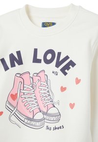 Witte sweatshirt met een grafischeafbeelding van roze hoge sneakers met witte veters, vergezeld van rode harten en de tekst "VERLIEFD op deze schoenen."