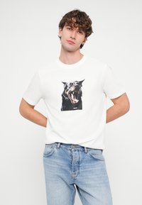 Balts kokvilnas T-krekls ar lielu nikna suņa attēlu, zem kura ir uzraksts „BOSS”. Valkāts ar ziliem džinsiem.