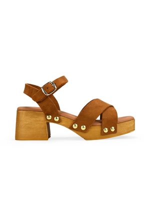 Sandalia de ante marrón con tacón bloque de madera, correa ajustable en el tobillo con hebilla plateada y tachuelas decorativas doradas en las correas.