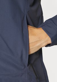 Marineblaues Jackett mit einer seitlichen Tasche und verstellbarem Kordelzug, gefertigt aus glattem, strapazierfähigem Stoff, das auf Passform und Funktionalität ausgerichtet ist.