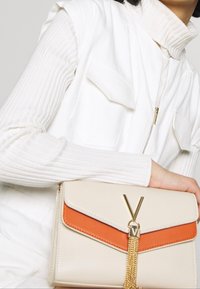 Beige a oranžová kabelka s klopou, zdobená zlatým V-čkovým akcentem a detailní řetízkou. Vyrobena z hladkého syntetického materiálu.