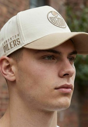 Junger Mann trägt beige Edmonton Oilers Mütze, schaut nach vorne, unscharfe Ziegelwand und Grünflächen im Hintergrund.