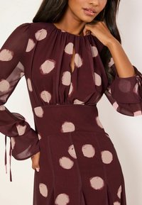 Burgundia punane kleit pikkade, läbipaistvate varrukatega, avausega kaeluses ja suurte kreemide täpiliste täppidega. Materjal paistab kerge ja sile, voolava disainiga.