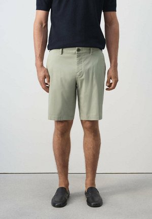 Uomo che indossa pantaloncini verde chiaro fino al ginocchio, mocassini neri e una camicia nera a maniche corte, in piedi su un pavimento grigio.