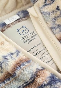 Blød, multifarvet babyjakke åbnet for at vise indvendig label med bjørnelogo og plejeinstruktioner på cremefarvet stof.