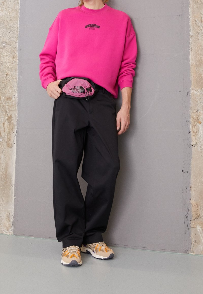 Personne portant un sweat-shirt rose vif, un pantalon noir, des baskets beiges et orange, et un sac banane rose et noir The North Face, debout contre un mur gris.