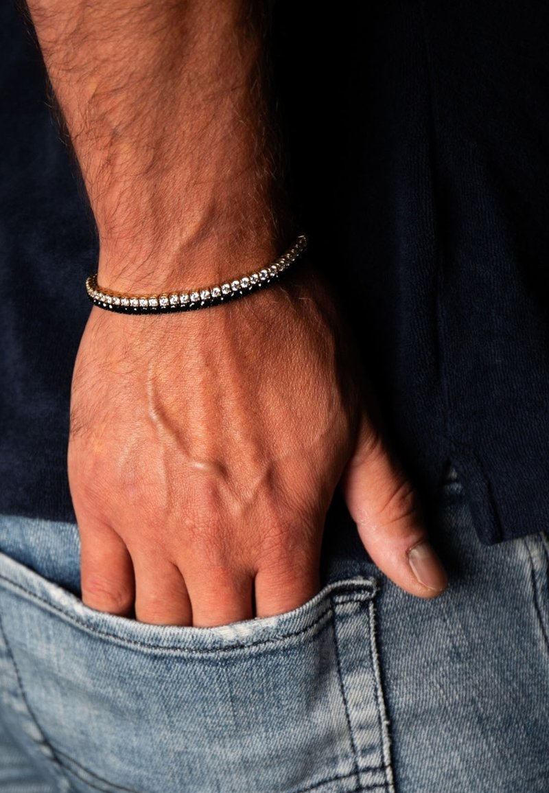 Bracciale con perline alternate in metallo argentato e nero, con un accento dorato, indossato su una mano appoggiata su un tessuto di denim.
