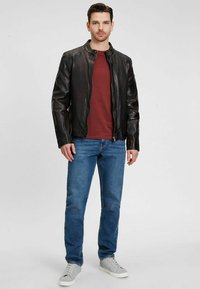 Schwarze Lederjacke mit Stehkragen, getragen über einem bordeauxfarbenen Hemd. Kombiniert mit blauen Jeans und grauen Sneakers. Glatte Textur, enganliegendes Design.