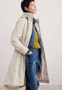 Lichtbeige lange jas met een hoge kraag, over een donkere spijkerblouse en een mosterdgele trui. Gecombineerd met blauwe spijkerbroek.