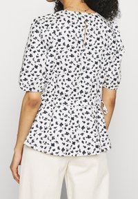 Hvid blomstret bluse med korte puffærmer, der har marineblå blomstermønster, en peplum talje og en nøglehulsryg med knaplukning.
