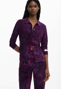 Chemise à fleurs violettes avec un design à boutons, dotée d'un col et de manches longues. Le tissu a une texture douce et un éclat subtil.