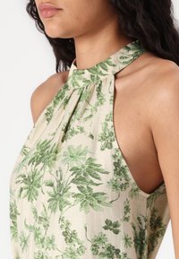Lahki halter top v bež tkanini z zelenim cvetličnim vzorcem. Ima naguban izrez in odprte strani za sproščen fit.