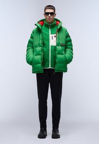 Grön puffjacka med huva, med en röd foder, bärs över en grön fleece och en vit zip-up jacka. Svarta byxor och solglasögon kompletterar looken.