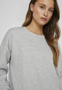 Pilkas džemperis suapvalintu kaklu ir ilgomis rankovėmis, priekyje išsiuvinėtu užrašu "ATELIER" šviesiai žalia spalva. Minkšta tekstūra.