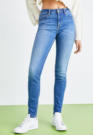 Femme portant un jean skinny bleu clair et des baskets blanches, debout sur un sol vert avec un arrière-plan blanc.