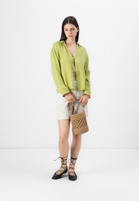 Limoengroene, lichtgewicht blouse met een V-hals en strikdetail, gecombineerd met beige shorts en een geweven bruine handtas. Zwarte veterschoenen.