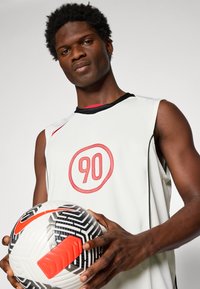 Utøver i ermeløs hvit sportstrøye med "90" i rødt, holder en hvit, svart og rød Nike fotball.