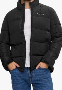 Abrigo acolchado negro con cuello alto, cierre de cremallera y diseño acolchado. Presenta una marca en el pecho y puños elásticos. Combinado con jeans azules.