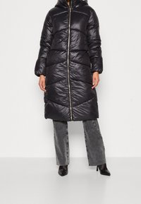 Manteau long matelassé en noir brillant avec une capuche, présentant un design matelassé diagonal, une fermeture éclair avant et des poignets côtelés ajustés.