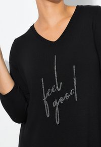 Schwarzes Langarm-T-Shirt mit V-Ausschnitt, das mit einem silbernen Strassschriftzug "feel good" gestaltet ist. Weicher Stoff.