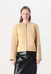 Veste en cuir brun avec fermeture éclair sur le devant, col rond et manches larges, associée à une jupe en cuir noire. Texture lisse, design minimal.