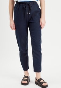 Pantalons en lin léger bleu marine avec une taille élastique et un cordon de serrage. Conception de jambe fuselée avec une texture subtile. Portés avec des sandales noires.