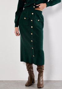 Jupe midi en maille verte à coupe droite, dotée de boutons dorés et de deux poches avant. Associée à des bottes hautes marron.