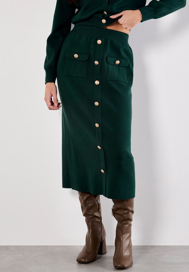 Jupe midi en maille verte à coupe droite, dotée de boutons dorés et de deux poches avant. Associée à des bottes hautes marron.