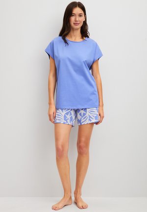 Frau steht barfuß vor weißer Wand, trägt ein lockeres hellblaues Oberteil und blaue Shorts mit weißem tropischem Blatt- und Seesternmuster.