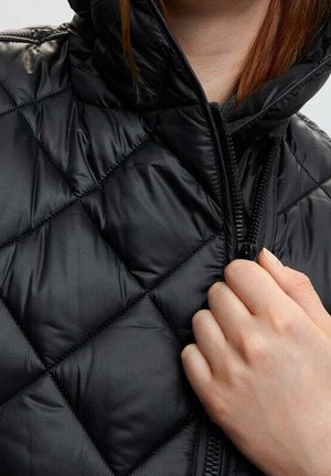 Mano che tiene la zip di una giacca nera trapuntata con collo alto, mostrando la texture lucida del tessuto e il motivo delle cuciture.